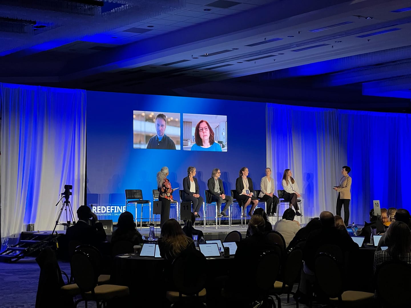 A seven person speaking panel at an event, two of which are on a screen. An example of a hybrid av event.